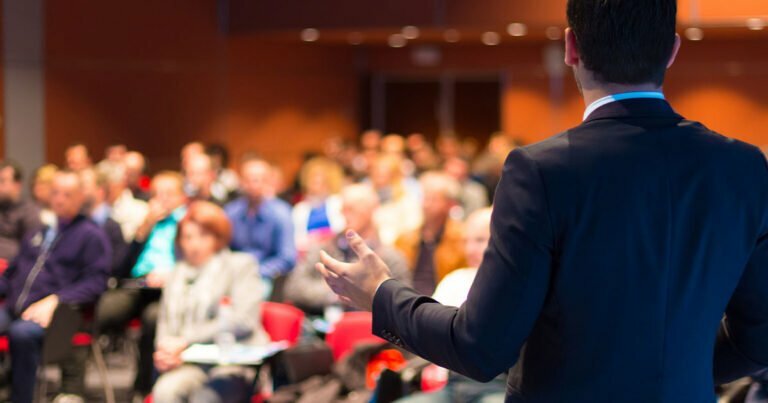 The back of a man on stage giving a speech in a packed event room