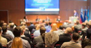 An out of focus pack event room where people are seated watching people talk