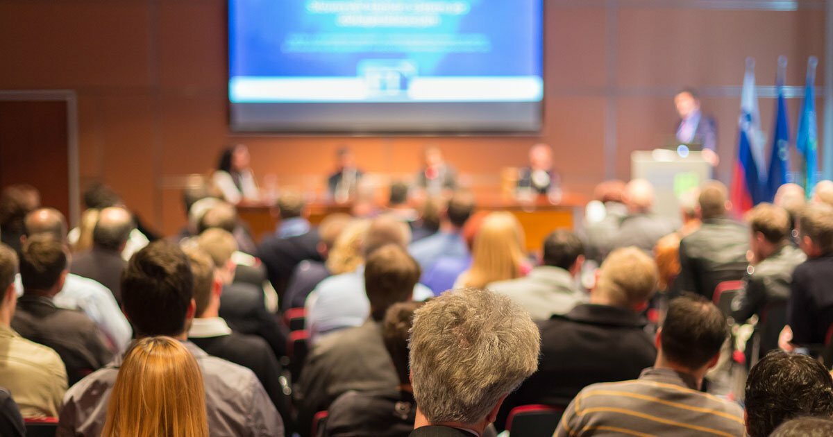 An out of focus pack event room where people are seated watching people talk