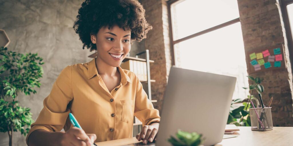 Happy lady in office taking notes while looking at laptop